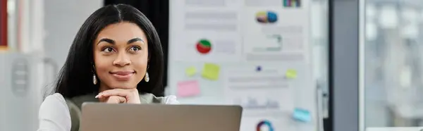 stock image A poised woman reflecting while using her laptop in a vibrant workspace.