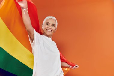 Olgun bir kadın neşeli bir şekilde rengarenk bir arka planda gökkuşağı bayrağı sallıyor..