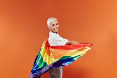 A mature woman joyfully wraps herself in a rainbow flag. clipart