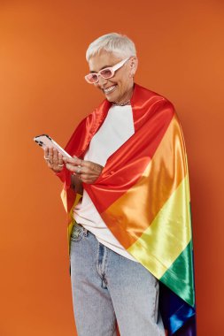 Olgun bir kadın, gökkuşağı bayrağına sarılmış telefonunu tutarken sıcak bir şekilde gülümsüyor..