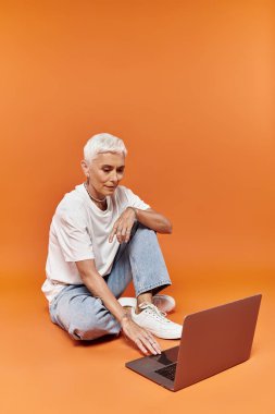 Mature woman with short hair interacts with a laptop in a vibrant setting. clipart