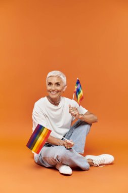 Olgun bir kadın parlak turuncu bir arkaplanda otururken elinde gökkuşağı bayrakları tutarken sevinç içinde.