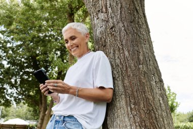 Olgun bir kadın mesajlaşırken gülümser, açık hava ve güneşin tadını çıkarır..