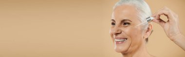 A mature woman smiles warmly while applying skincare.