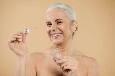 Olgun, kısa saçlı bir kadın neşeyle serum kullanıyor..