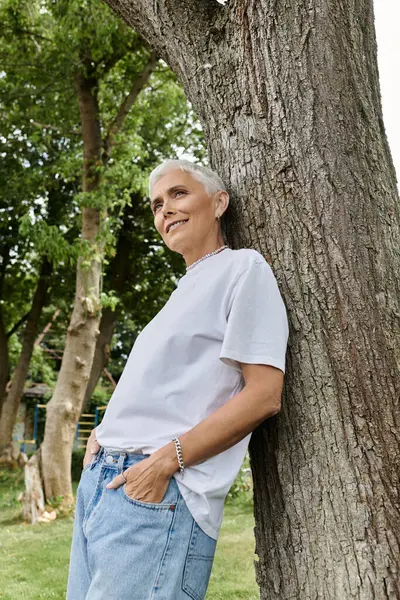 stock image A mature woman with short hair leans against a tree, embracing the natural beauty around her.