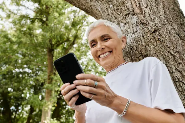 Stock image A mature woman smiles as she texts under the shade of a tree.