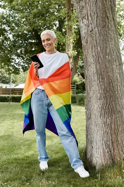 Kısa saçlı olgun bir kadın gururla durur gökkuşağı bayrağına sarılmış gülümserken.