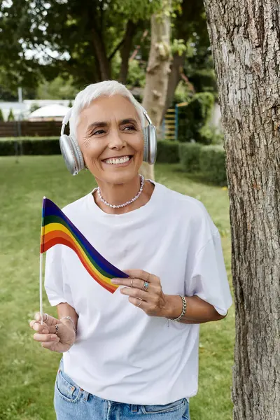 stock image A smiling woman holds a pride flag while enjoying music in a sunny park.