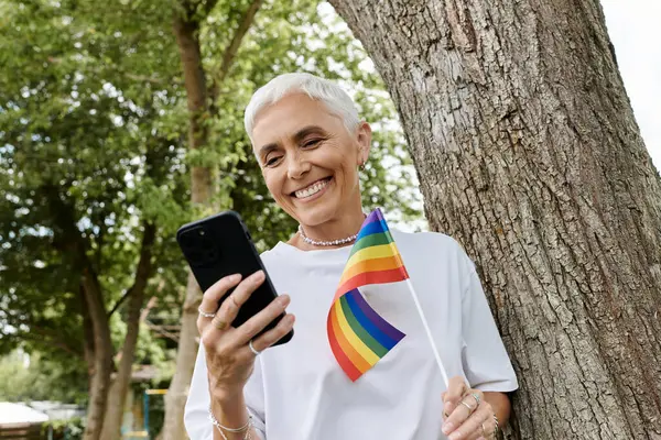 Havalı bir kadın gülümser, parkta telefonunu kontrol ederken gökkuşağı bayrağı sallar..