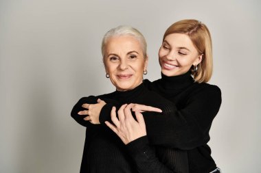 A mature woman smiles while her daughter embraces her warmly from behind. clipart