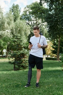 Dövmeli genç bir adam akıllı telefonunu kullanarak yeşil bir parkta yürüyor.