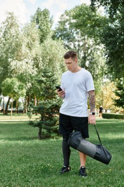 Dövmeli genç bir adam açık havada akıllı telefonunu kullanarak yeşil bir parkta yürüyor.