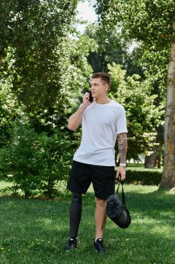 Dövmeli genç bir adam yeşil bir parkta yürüyor, dışarıda akıllı telefonuyla konuşuyor.