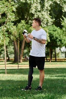 Dövmeleri ve protez bacağı olan genç bir adam canlı yeşil bir parkta dururken müzik dinliyor..