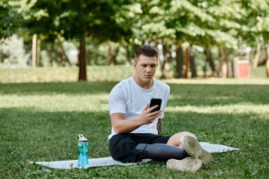 Dövmeli protez bacaklı genç adam spor minderinde oturuyor ve parkta akıllı telefon kullanıyor.