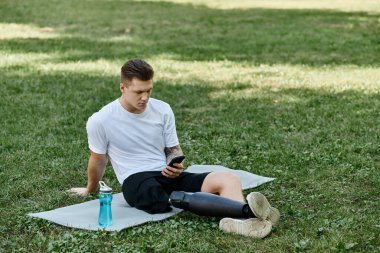Dövmeli protez bacaklı genç adam spor minderinde oturuyor ve parkta akıllı telefon kullanıyor.