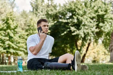 Dövmeli, protez bacaklı genç adam spor minderinde oturuyor ve telefon görüşmesi yapıyor, parkta akıllı telefon kullanıyor.