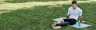 Dövmeli, protez bacaklı genç adam spor paspasında oturuyor ve yeşil parkta telefon kullanıyor.