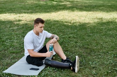 Dövmeli genç adam protez bacaklı su şişesi tutuyor ve parkta mindere oturuyor, su ve spor yapıyor.