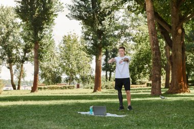 Tattooed young man with artificial leg working out in green park, engaging in an online fitness lesson clipart