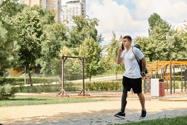 Dövmeli genç bir adam yeşil bir parkta yürüyor, telefonuyla konuşuyor.