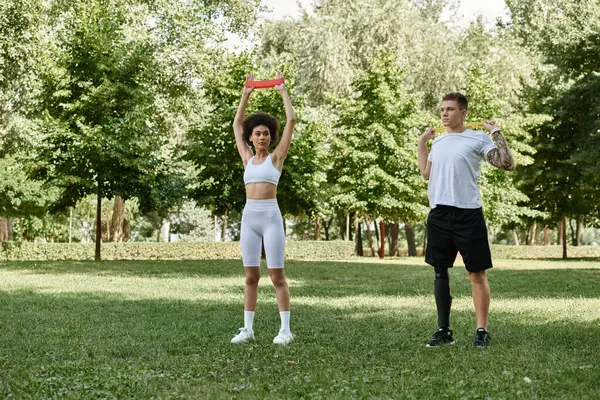 stock image A young woman and a tattooed man exercise in a lush park, showcasing fitness and friendship.