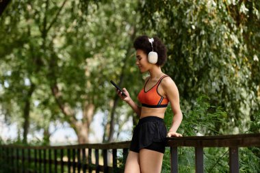 A woman standing on a bridge, listening music in headphones surrounded by vibrant trees and fresh air. clipart