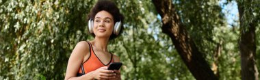 Genç bir kadın akıllı telefon kullanıyor ve parkta kablosuz kulaklıkla müzik dinliyor.