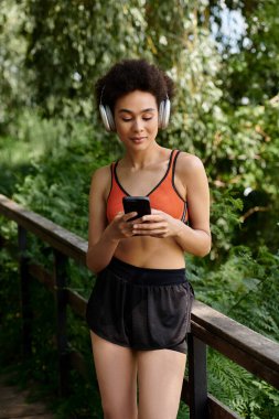Akıllı telefon kullanan ve kulaklıkla müzik dinleyen genç bir kadın. Etrafı yeşil ağaçlar ve temiz havayla çevrili..