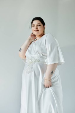 Göz kamaştırıcı, büyük beden bir gelin beyaz gelinliğini ve zarif küpelerini sessiz arka planda sergileyerek zarifçe poz verir..