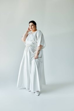 Büyüleyici bir gelin, zarif beyaz elbisesini ve parlak küpelerini soluk gri arka planda sergiliyor..