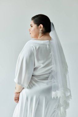 Güzel, genç bir gelin, beyaz elbisesi ve göz kamaştırıcı küpeleriyle sade, gri bir arka plana ışık saçar..