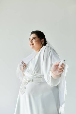 Güzel, genç bir gelin, beyaz bir elbiseyle ışıldıyor. Küpeleri var. Gri bir arkaplanda neşe saçıyor..