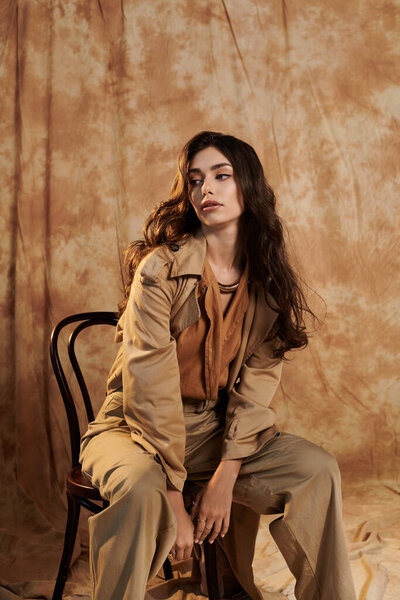 A young woman highlights her autumn style, seated thoughtfully in a warm-toned studio, embodying elegance and comfort.