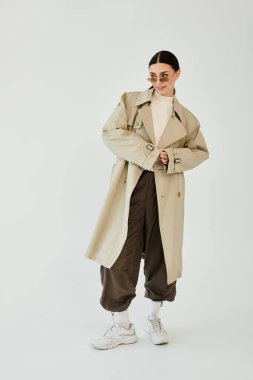 Genç bir kadın, şık bir sonbahar kıyafetinin içinde, bir stüdyo arka planında kendine güvenle poz verir..