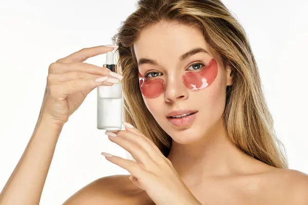 stock image A young woman showcases her skincare routine, holding a serum while wearing eye patches.