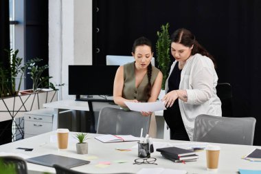 İki profesyonel kadın, bitkilerle dolu şık bir ofis çalışma alanında evrak işlerini tartışıyor..