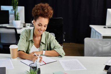 Profesyonel bir kadın, modern ofis çalışma alanında notlar alırken gülümser..