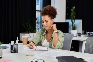 A professional woman deeply concentrating as she takes notes at her stylish office desk. clipart