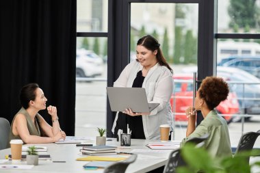 A confident businesswoman presents ideas on her laptop to engaged colleagues in an office. clipart