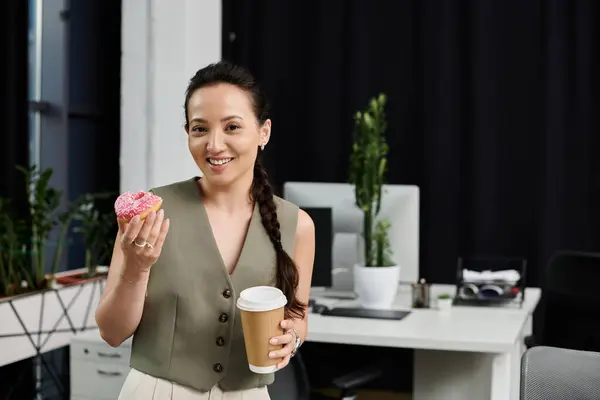 stock image A professional woman smiles radiantly while holding a coffee and a bright donut in her office.