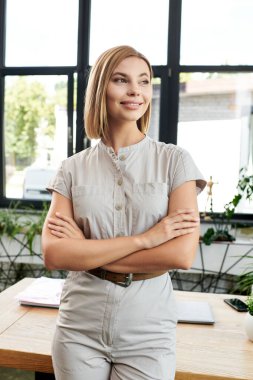 Kendine güvenen genç bir profesyonel kollarını kavuşturmuş, hareketli bir ofis ortamında gülümsüyor..