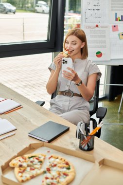 Genç bir kadın ofis masasında akıllı telefonunu kontrol ederken bir parça pizza yer..