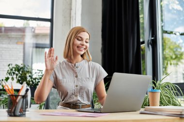 A cheerful professional waves at laptop in a sunny workspace filled with greenery. clipart