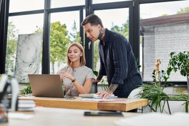 Two coworkers engage in a lively discussion while working on a laptop at their desk. clipart