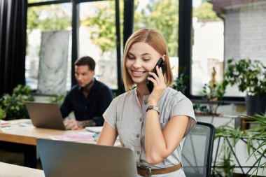 Neşeli genç bir kadın, parlak bir ofiste bilgisayarının başında çalışırken telefonla konuşur..