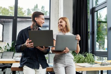 İki genç profesyonel modern bir ofiste dizüstü bilgisayarları tutarken projeleri neşeyle tartışıyor..