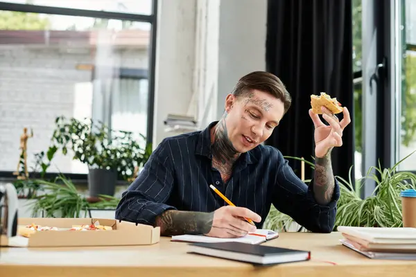 stock image A young man jots down ideas in a notebook with one hand while holding a pizza in the other.