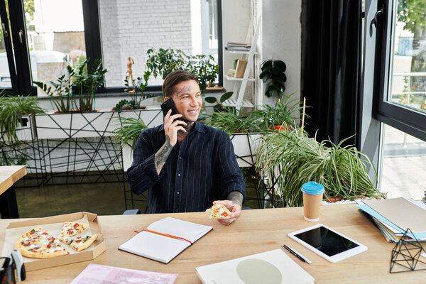 A cheerful worker talks on the phone, enjoying pizza at a vibrant and green workspace.